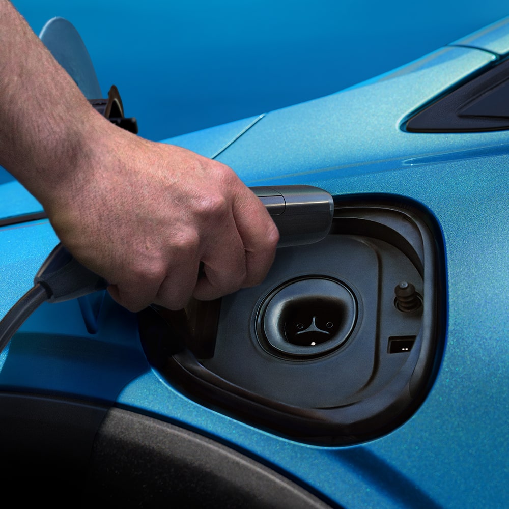 A Hand Plugs a Charging Cable into the Charging Port of a Light Blue 2027 Chevrolet Bolt.