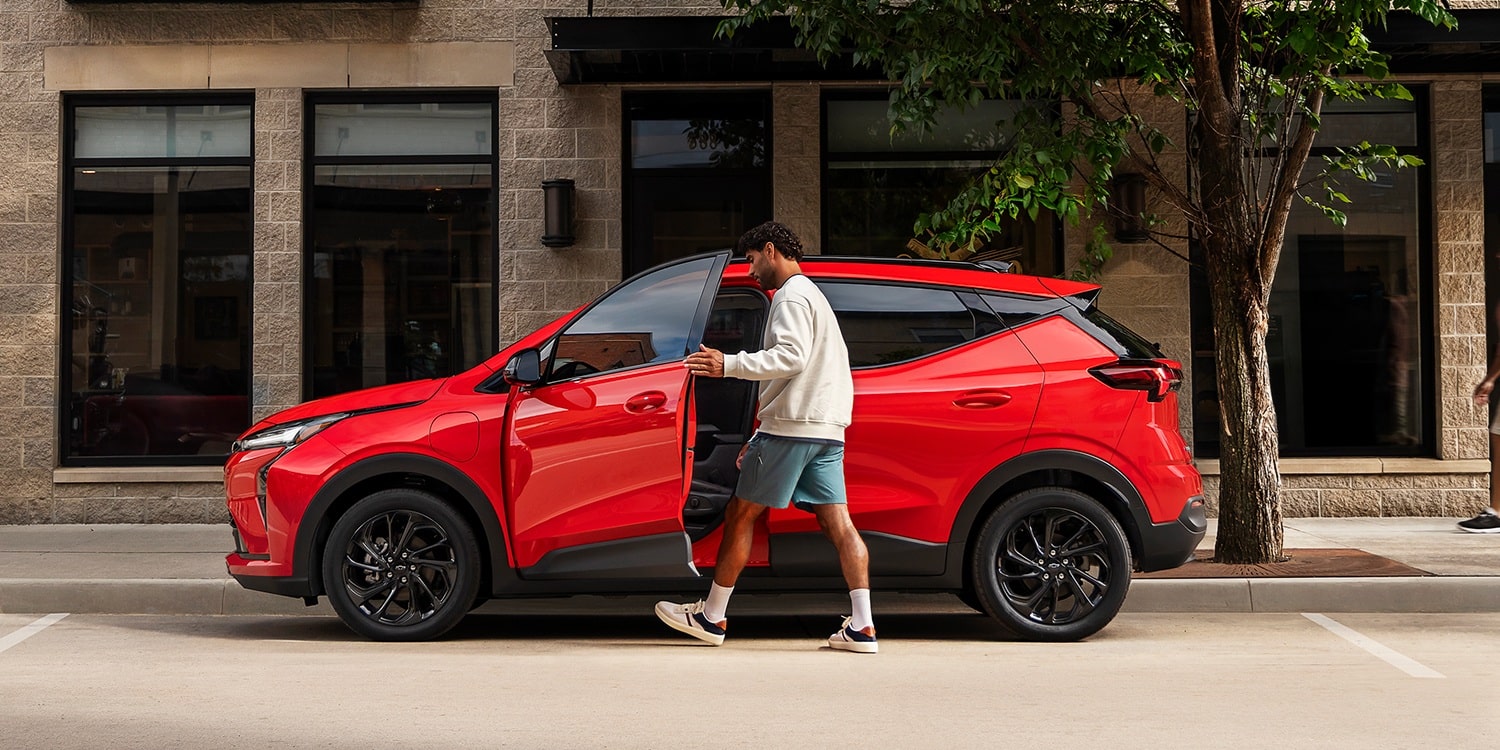 The Side View of a Red 2027 Chevrolet Bolt, with a Man Getting into the Driver's Side Door on a City Street.