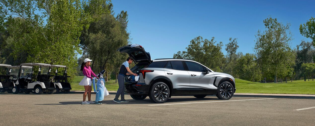 Two people loading golf equipment into a silver Chevrolet Blazer EV parked at a golf course.