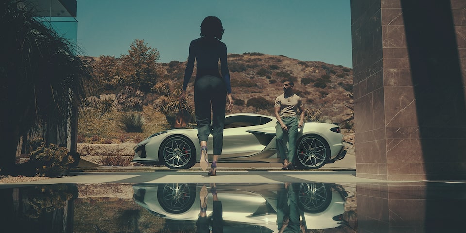 A man leans against his Corvette Z06 as a woman approaches with a mountain backdrop.