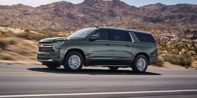 Exterior driver side view of the 2023 Chevrolet Suburban driving on a country road.