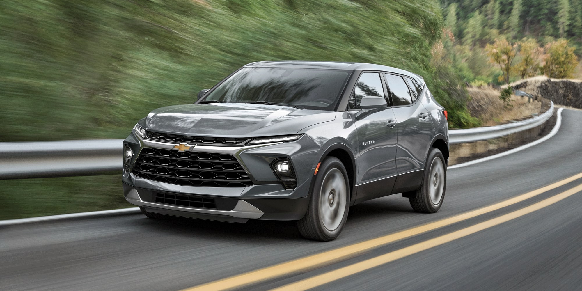 Un Chevrolet Blazer 2026 en gris sterling métallisé qui roule à toute allure sur une route bordée d’arbres.