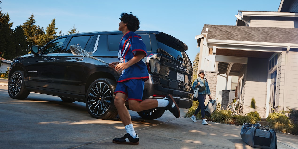 A Young Lacrosse Player Running past the 2026 Chevrolet Suburban Followed by His Mom Outside of Their Home.