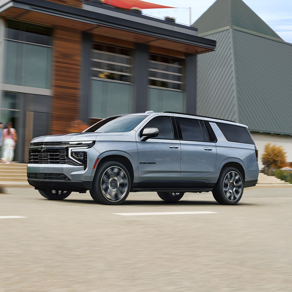 Side View of the 2026 Chevrolet Suburban SUV Driving Past a Modern Apartment Building.