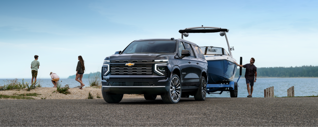 Un Chevrolet Suburban 2026 qui remorque un bateau devant un lac, avec des gens qui marchent à côté.
