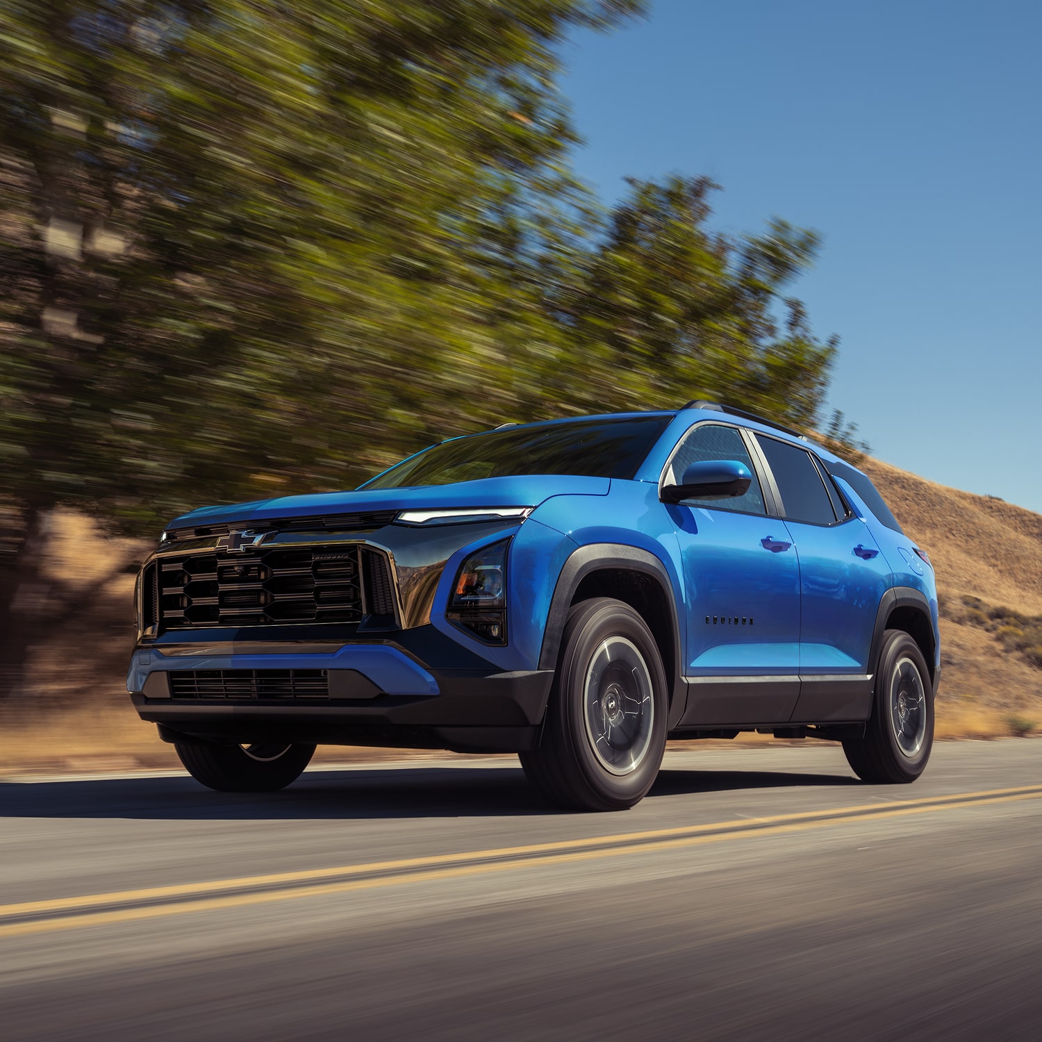 A 2027 Blue Chevrolet driving down the street line with bushes.