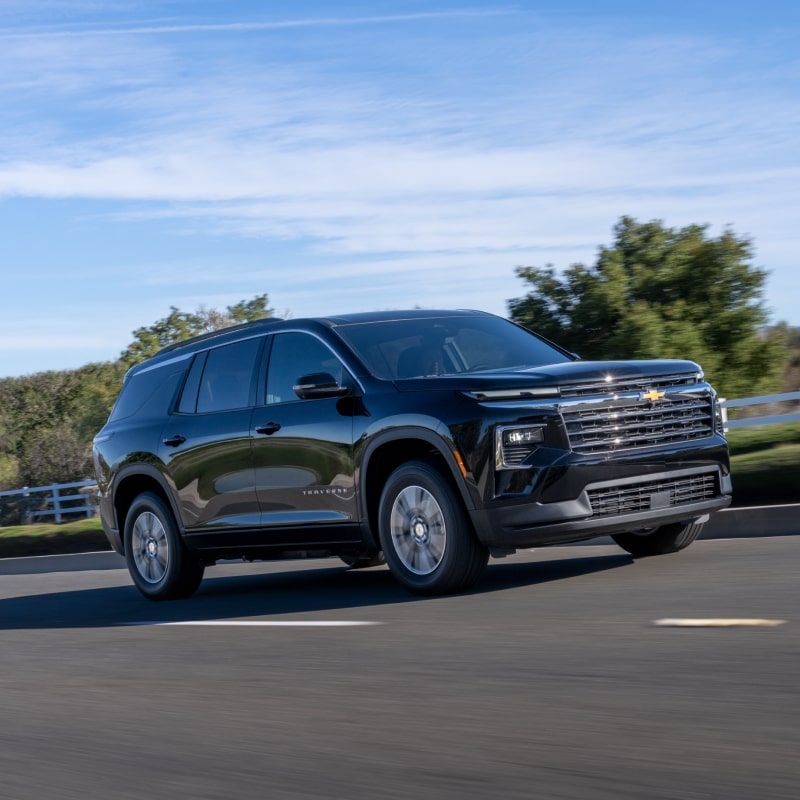 Vue latérale de la Chevrolet Traverse 2027 roulant sur une route bordée d’arbres.