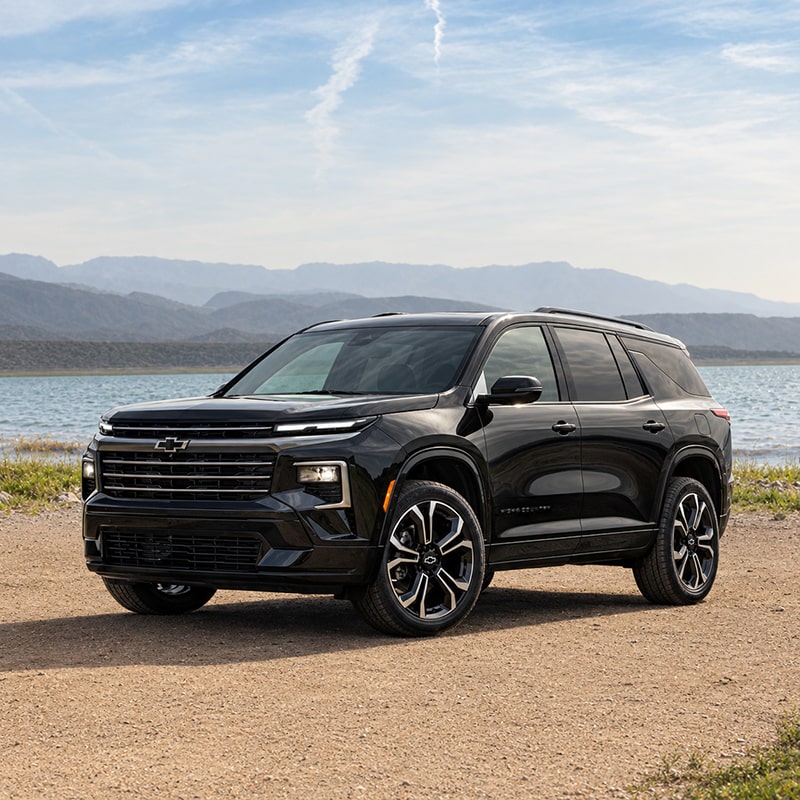 Side view of a 2027 Chevrolet Traverse parked on a lakeside dirt road under a lightly clouded sky.