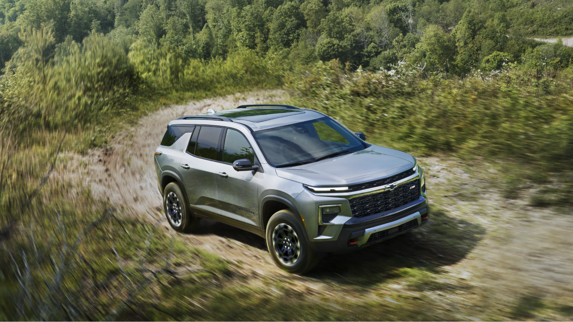 Une Chevrolet Traverse 2027 roulant seule sur une route bordée d’arbres.