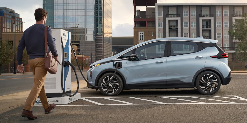 Man walking towards 2023 Bolt EUV plugged into charger in city.