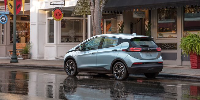 Exterior rear driver side view of the 2023 Bolt EV parked on a rain-soaked town street.