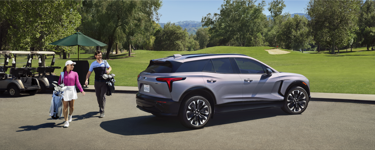 Two people with golf clubs walking past a Chevrolet Blazer EV parked at golf course.