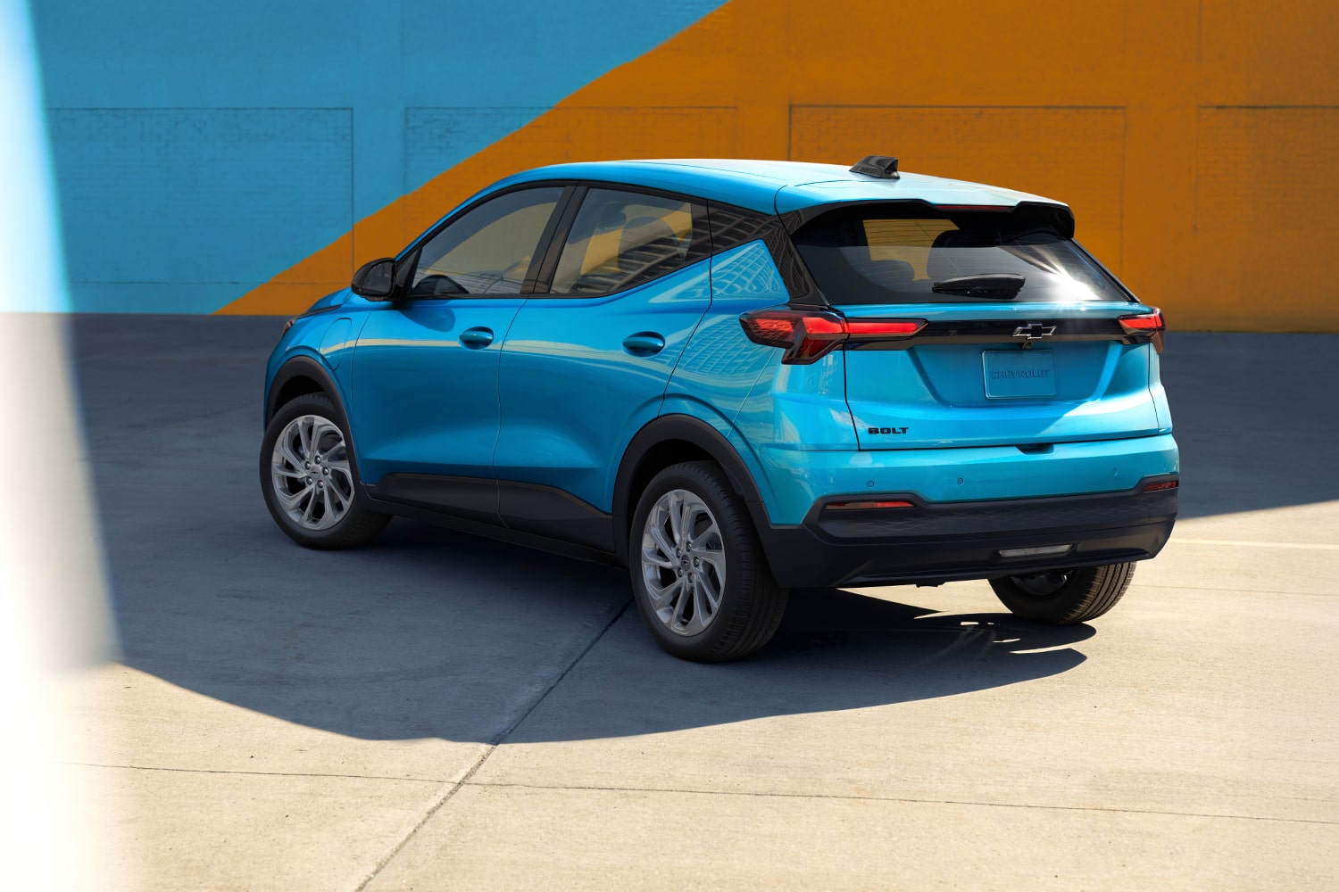 The Rear View of a Light Blue 2027 Chevrolet Bolt Shows the Vehicle Parked in Front of a Blue and Orange Wall, Highlighting the Updated Taillights and Design.