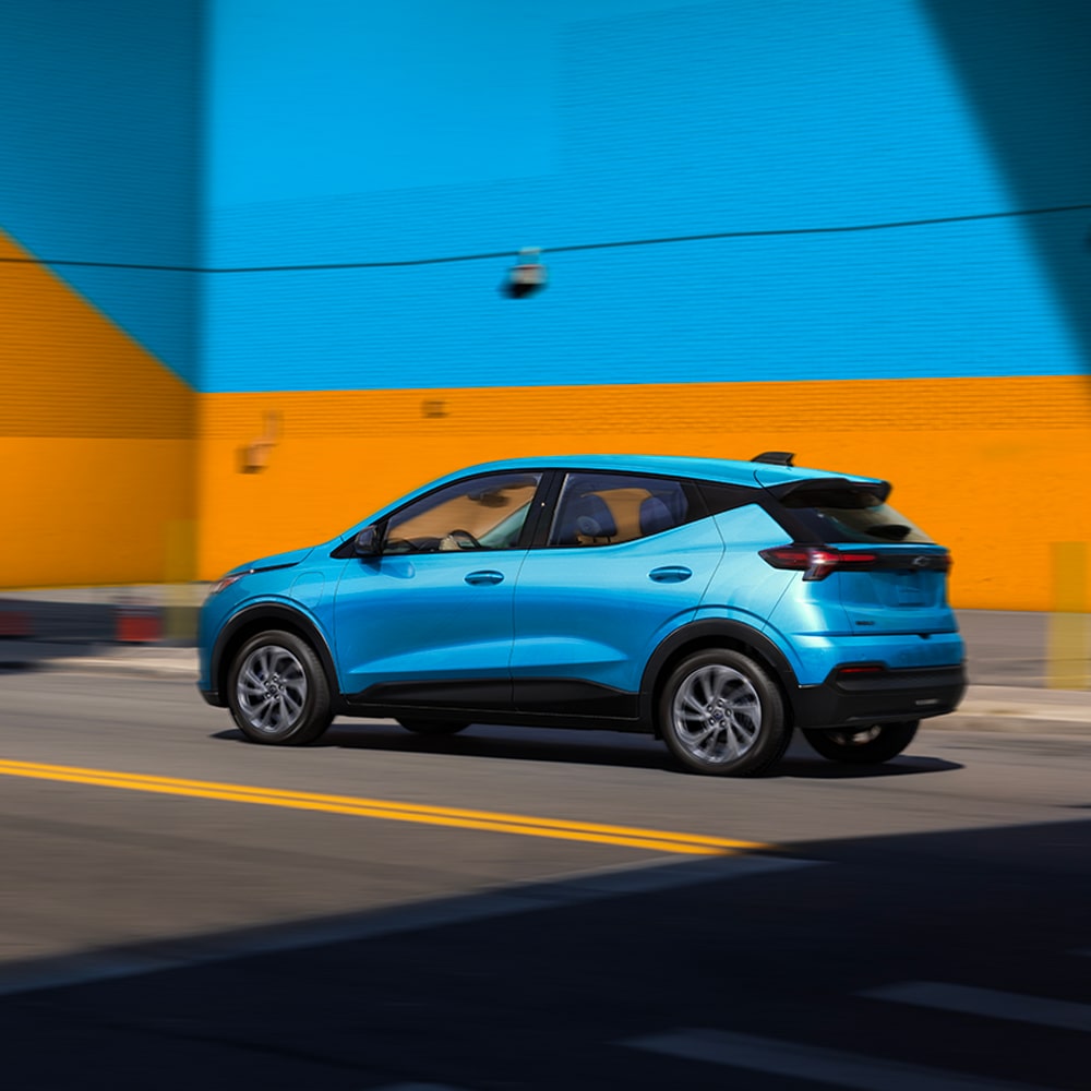 The Rear View of a Light Blue 2027 Chevrolet Bolt Shows the Vehicle Parked in Front of a Blue and Orange Wall, Highlighting the Updated Taillights and Design.