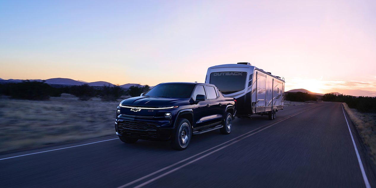 A black Chevrolet Silverado EV towing a trailer on the road at sunset.