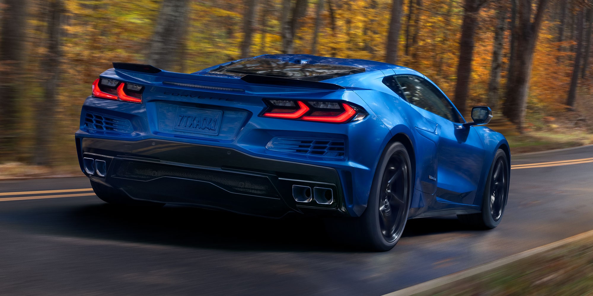 Rear View of a 2026 Chevrolet Corvette E-Ray Driving Down a Road Surrounded by Trees.