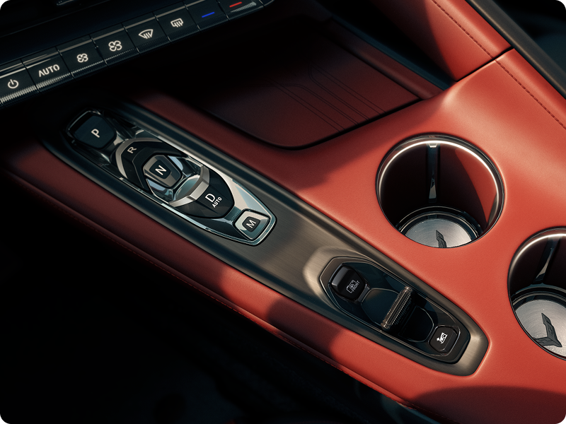 A Close‑Up View of the Center Console in a 2027 Chevrolet Corvette Grand Sport Shows Metallic Switch Controls Beside Two Circular Cupholders Set in Red Interior Trim.