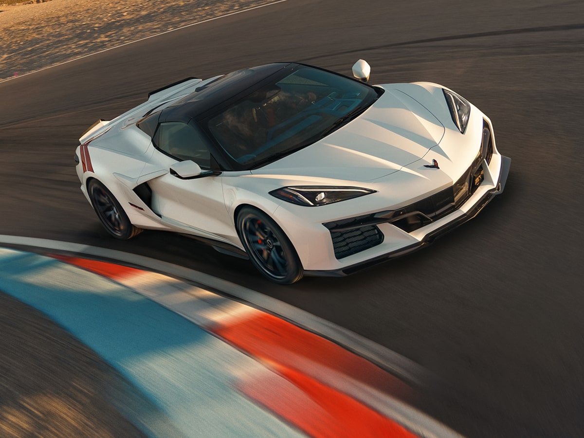 A White 2027 Chevrolet Corvette Grand Sport With Red Accent Stripes Leans Into a Curved Racetrack Section With Motion Blur Across the Painted Apex.