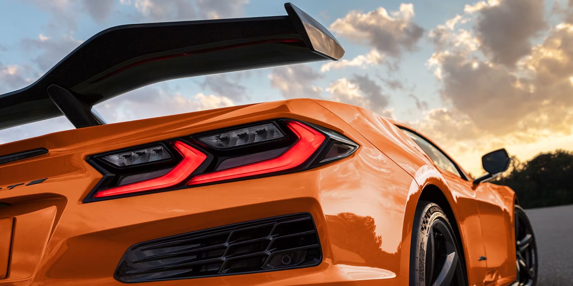 Gros plan sous un angle arrière d'une Chevrolet Corvette Z06 orange de 2027, dotée d'un grand aileron noir en fibre de carbone, sur fond de ciel au coucher du soleil.