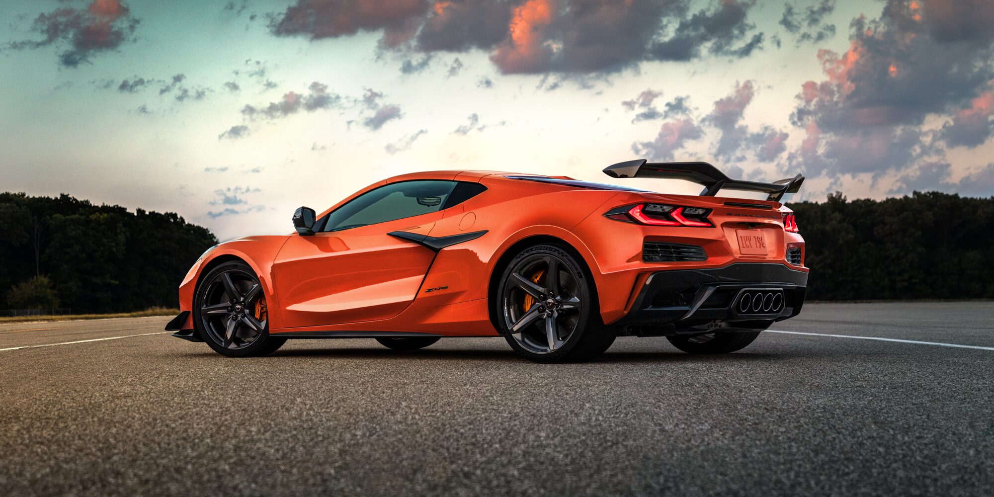 Vue en contre-plongée d'une Chevrolet Corvette Z06 orange (modèle 2027) garée sur une route dégagée, face à un magnifique coucher de soleil.