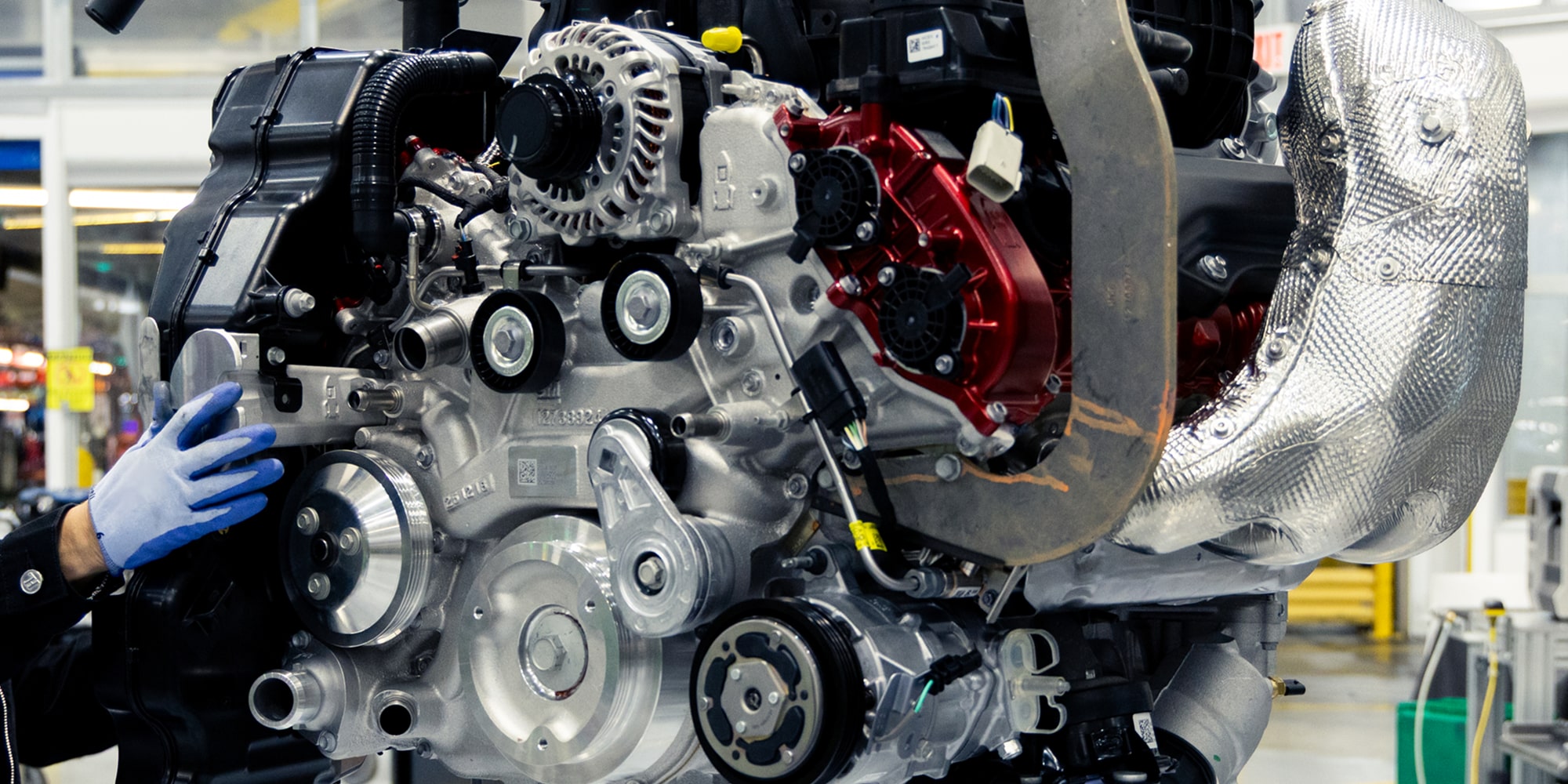 Un ouvrier ganté assemble un moteur Chevrolet avec des composants métalliques apparents et des boyaux sur une chaîne d’assemblage d’usine.