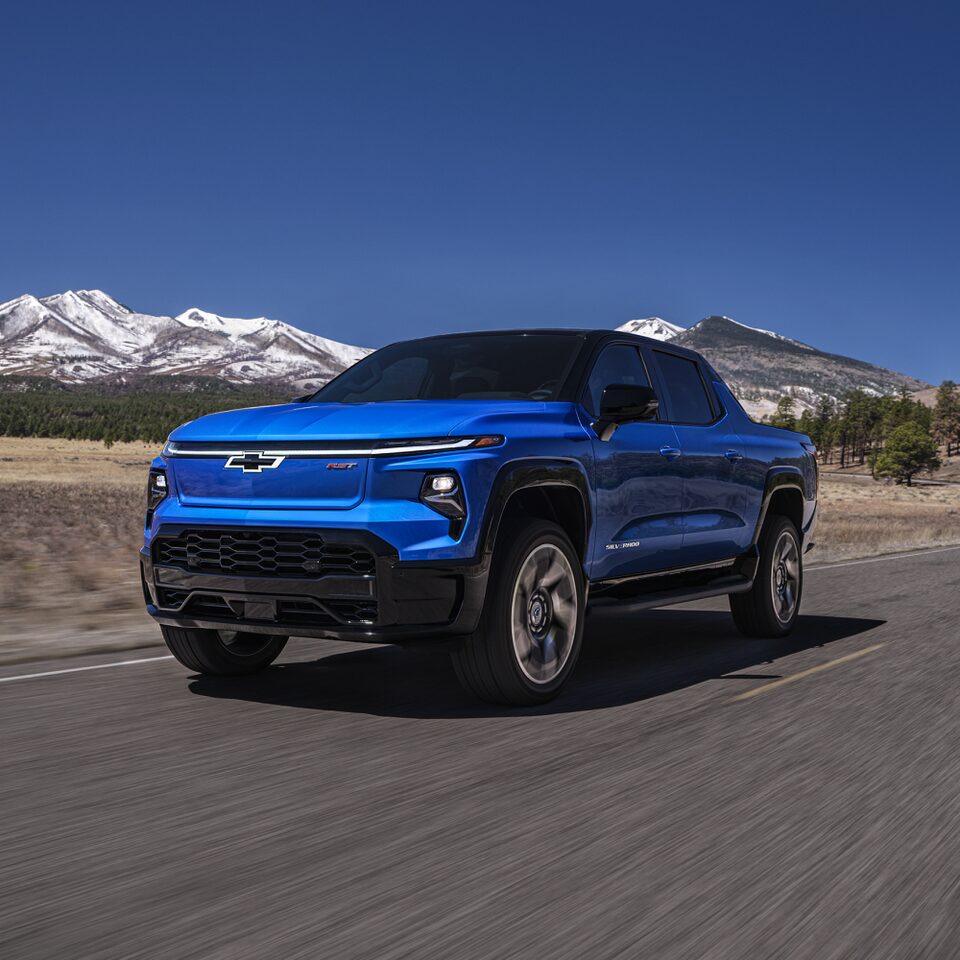 Three-quarters view of the front end of the 2024 Chevrolet Silverado EV driving along a forest-lined road.