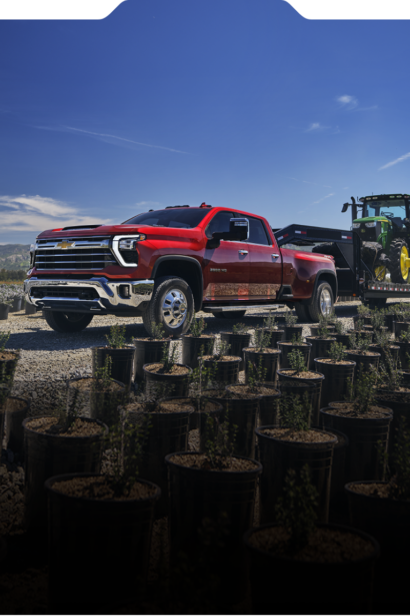 The new 2027 Chevrolet Silverado HD pickup truck towing a green tractor on a trailer through an agricultural nursery.