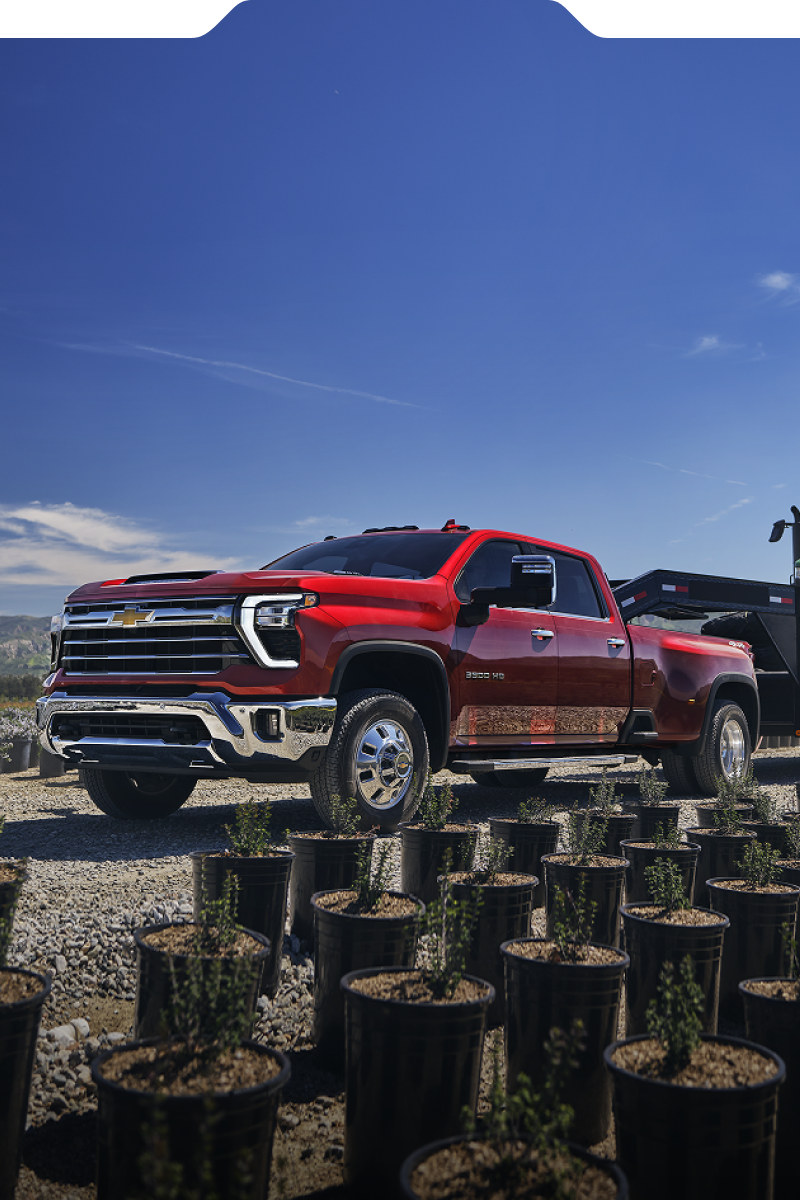 Le nouveau camion Chevrolet Silverado HD 2027 remorquant un tracteur vert sur une remorque à travers une pépinière agricole.