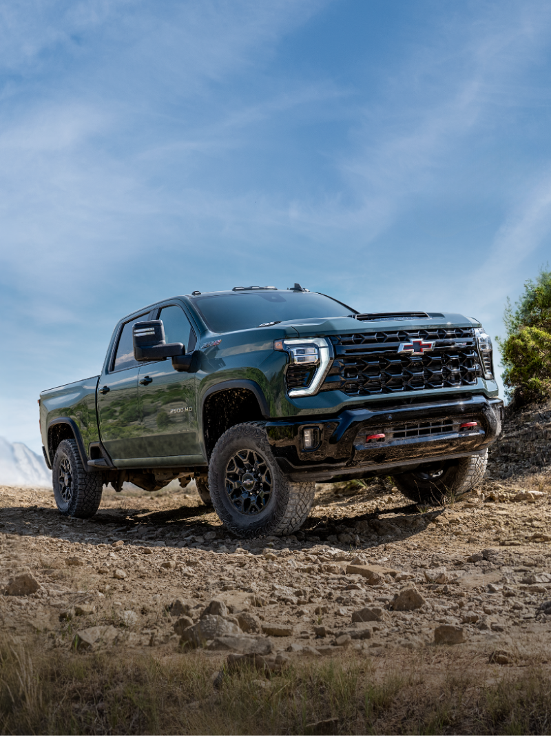 Vue frontale des trois quarts du nouveau pick-up Chevrolet Silverado HD 2027 roulant sur un sentier rocheux hors route, avec des montagnes et un ciel bleu en arrière-plan.