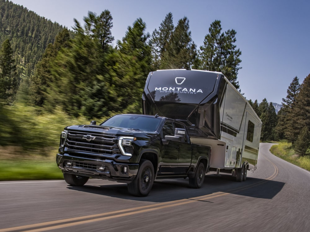 The new 2027 Chevrolet Silverado HD pickup truck towing a large RV trailer along a winding highway through a forested mountain landscape.