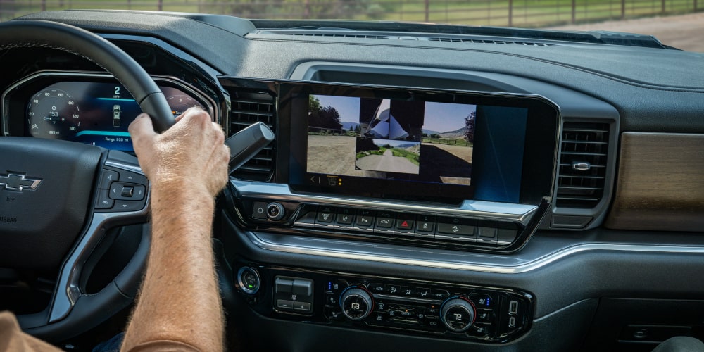 Interior view of a 2027 Chevrolet Silverado HD cabin showing a driver using the steering wheel while the center touchscreen displays a trailer camera monitoring system.