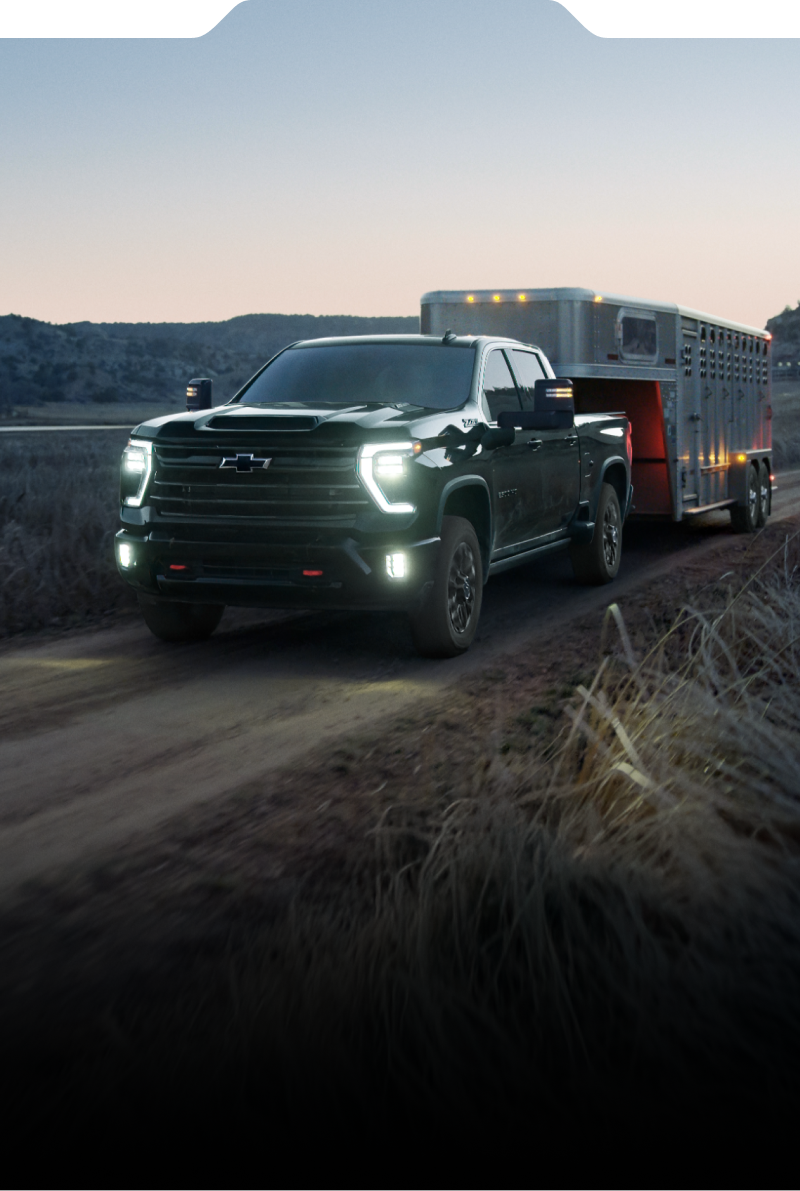 The new 2027 Chevrolet Silverado HD pickup truck towing a trailer along a rural dirt road at dusk.