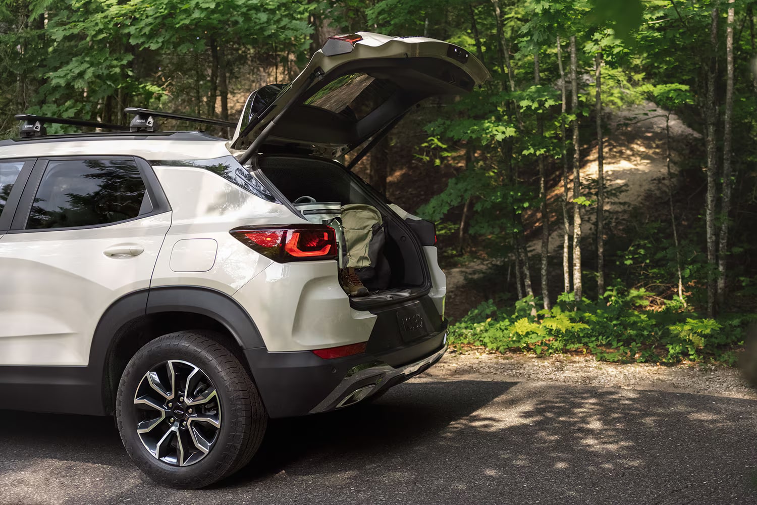 Side rear view of a white 2027 Chevrolet Trailblazer parked on a forest road with the liftgate open, showcasing the cargo area.