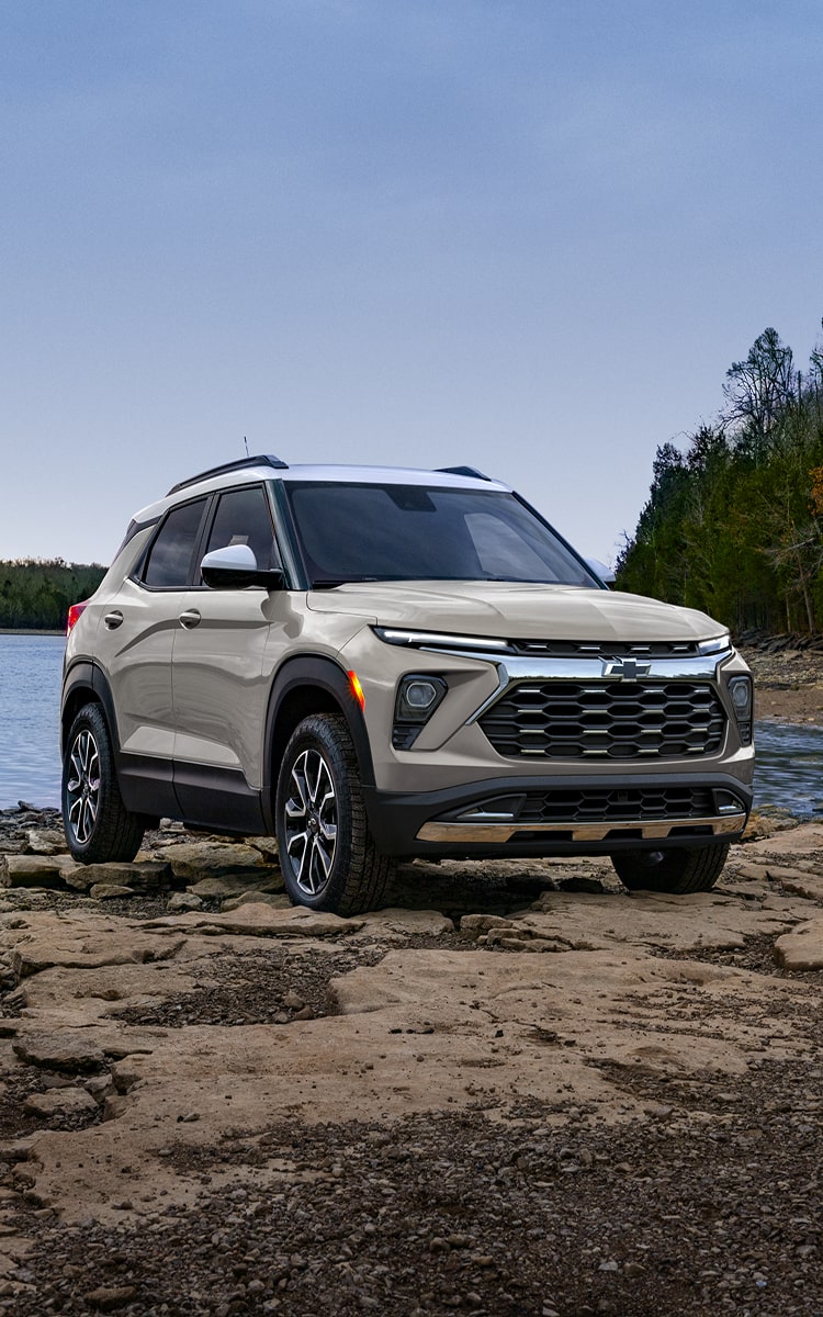 Side view of a 2027 Chevrolet Trailblazer driving along a paved road through a lush forest.