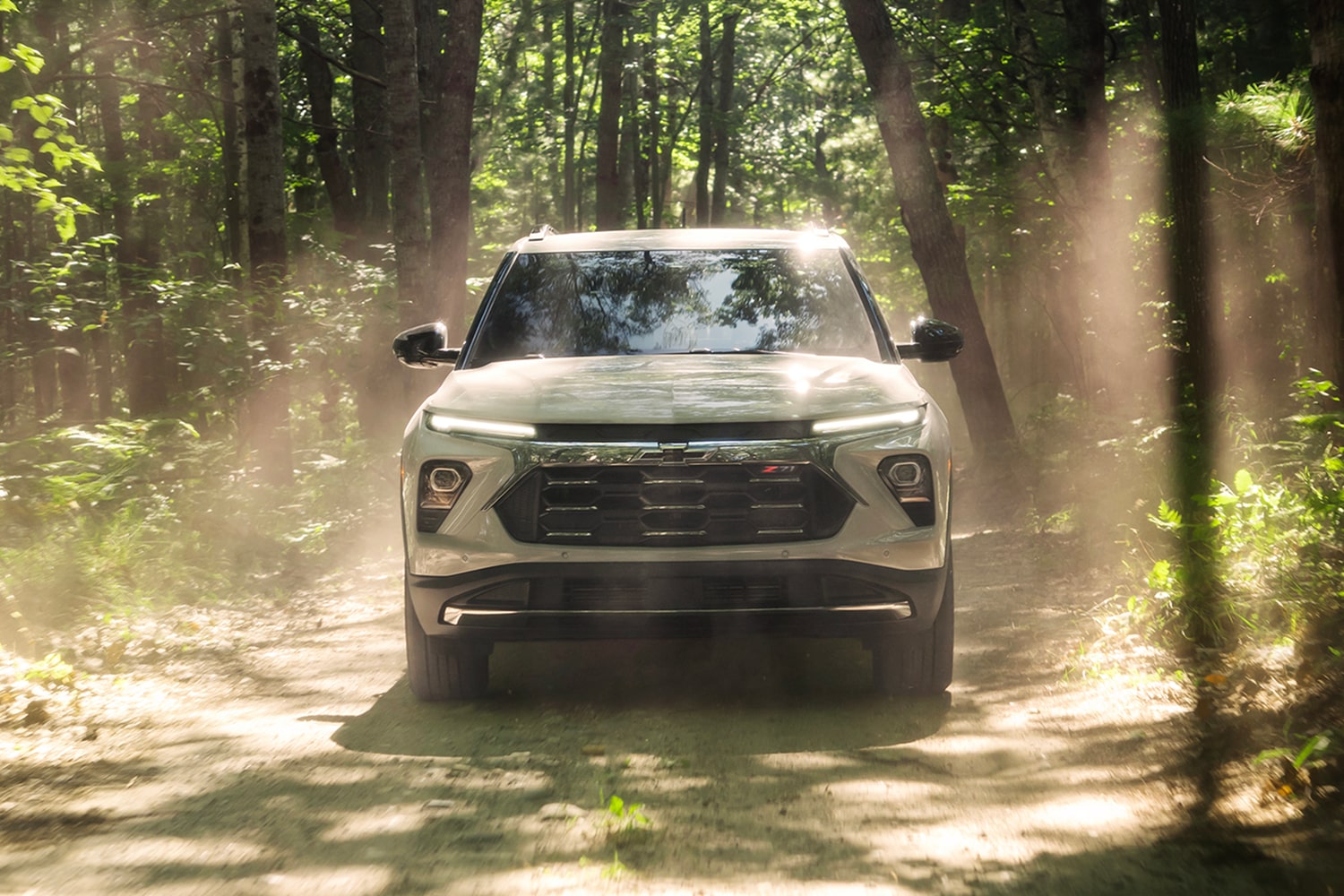 Front view of a 2027 Chevrolet Trailblazer driving on a wooded trail, surrounded by tall trees and sunlight filtering through the forest.