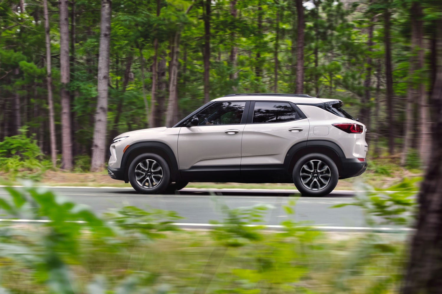 Side view of a 2027 Chevrolet Trailblazer driving along a paved road through a lush forest.