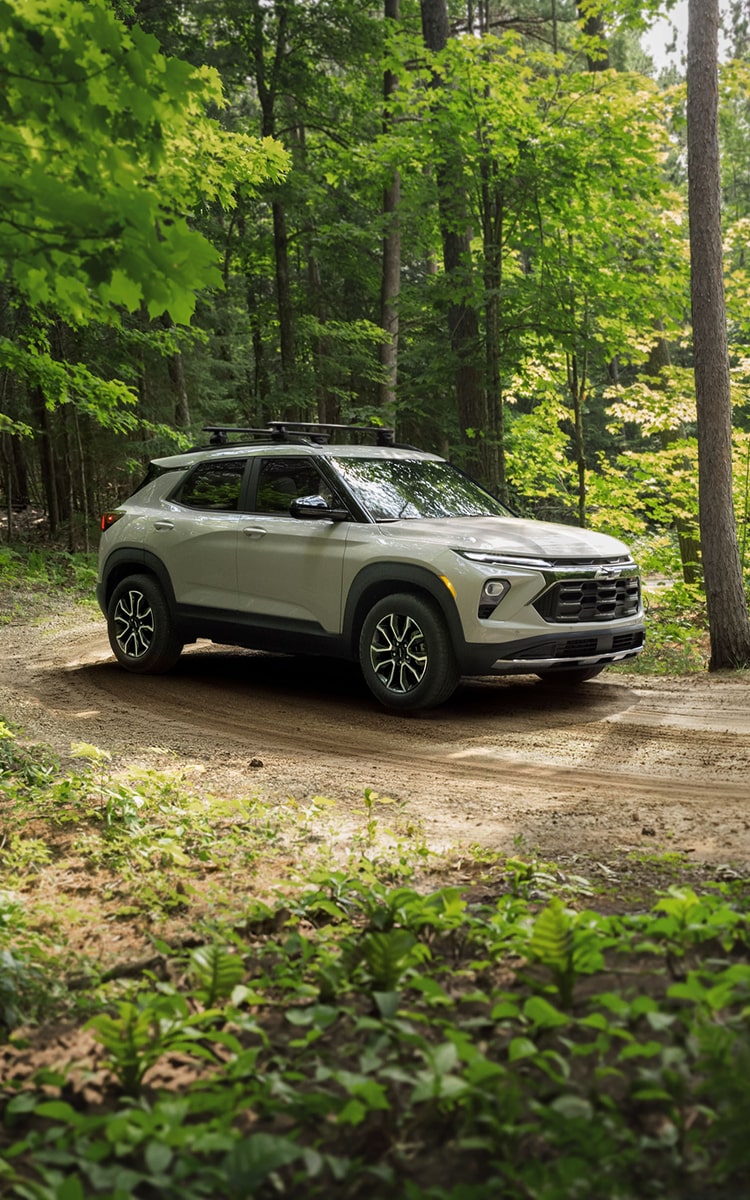A 2027 Chevrolet Trailblazer SUV driving on a dirt trail through a lush green forest, surrounded by tall trees and natural woodland scenery.