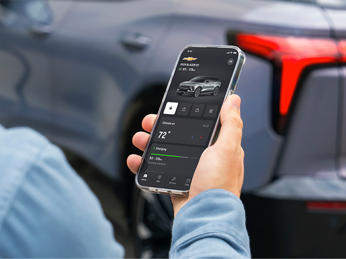 A hand holding a smartphone displaying the OnStar mobile app in front of a 2027 Chevrolet Trailblazer SUV.