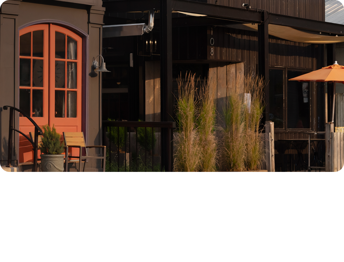 Close-up of a building exterior with warm orange-trimmed windows and doors, dark architectural accents, potted plants, and soft natural lighting.