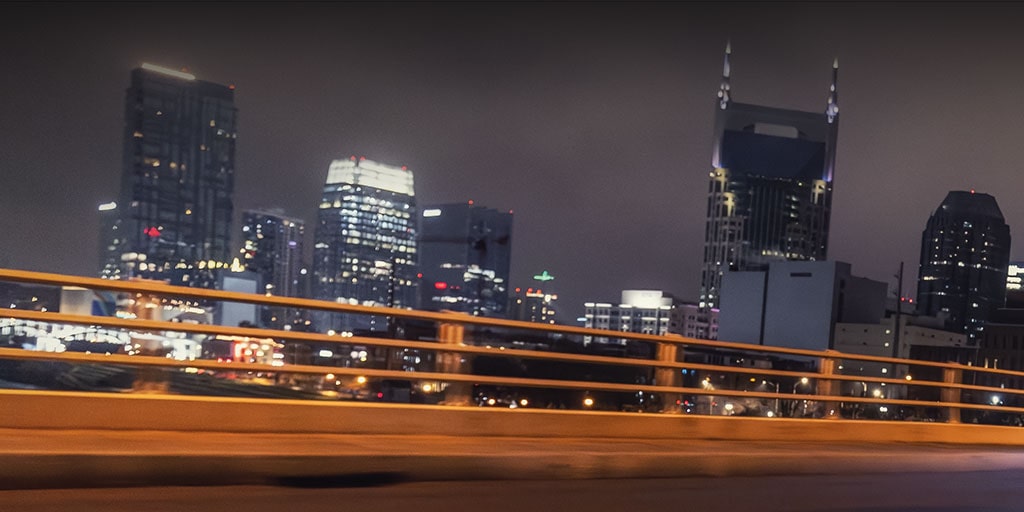 A city skyline at night with illuminated high-rise buildings and bridge railings in the foreground.
