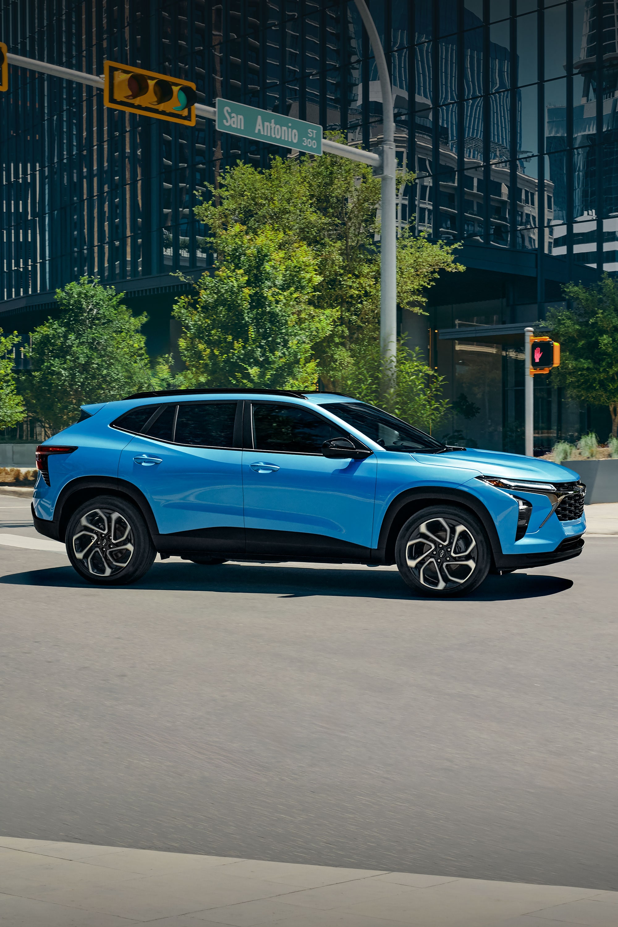 2027 Chevrolet Trax small SUV side profile driving through a modern downtown intersection with blue exterior paint, alloy wheels, and city buildings in the background.