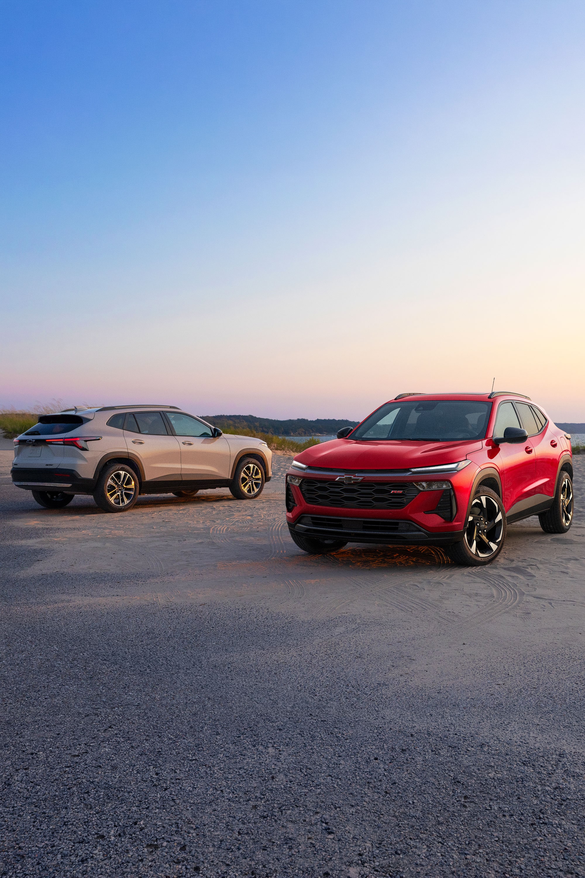 Red and white 2027 Chevrolet Trax small SUV models parked on a sandy coastal landscape at sunset, showcasing front and rear three-quarter exterior views.