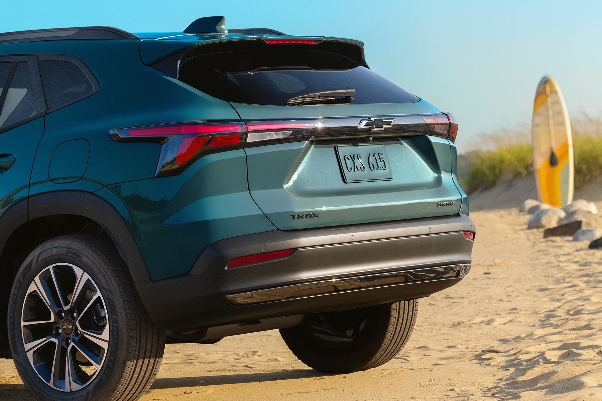 2027 Chevrolet Trax small SUV rear three-quarter view parked near a beach with LED taillights, alloy wheels, and a surfboard in the background.