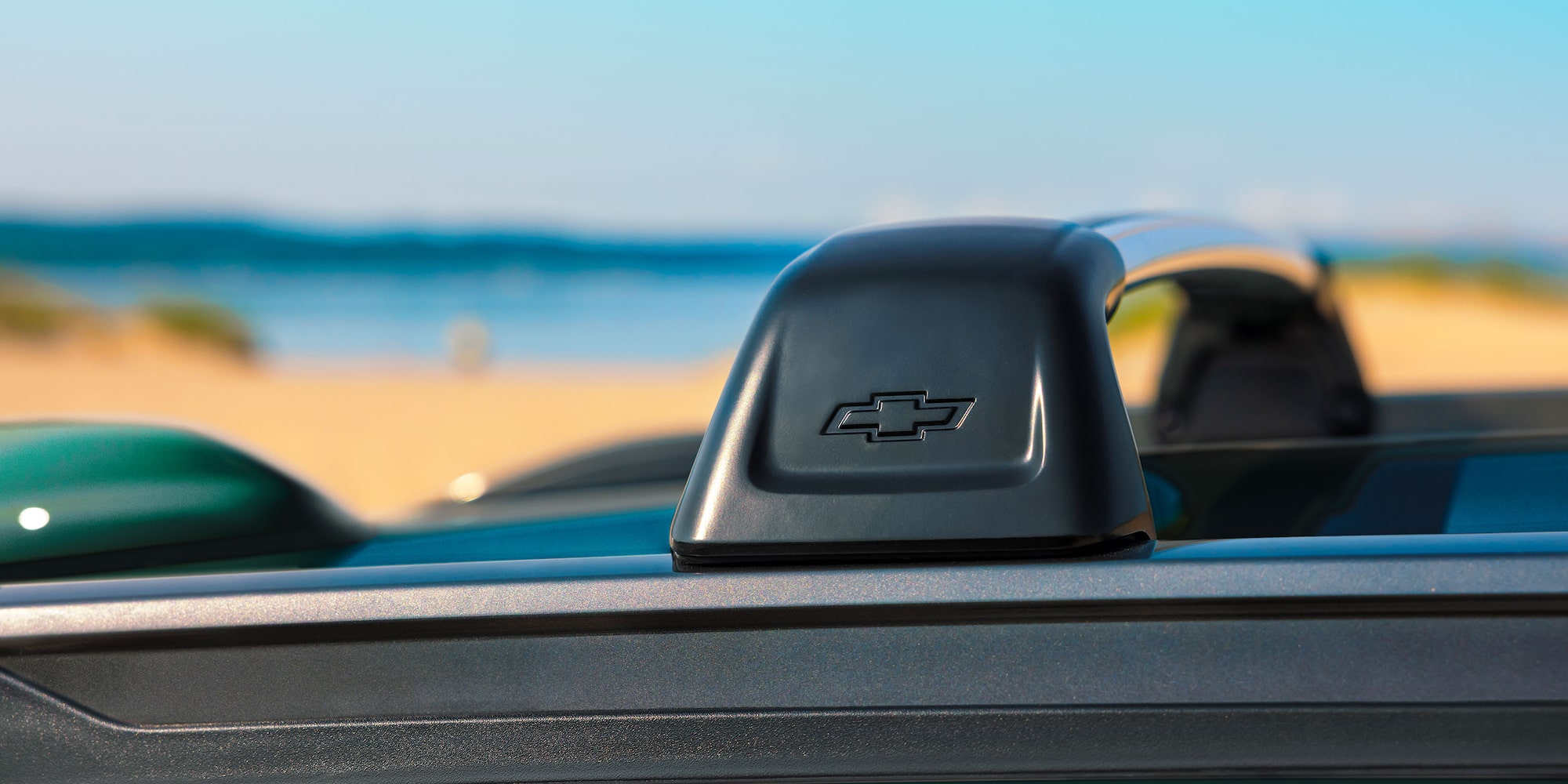 Close-up of the roof rail on the 2027 Chevrolet Trax small SUV, featuring the Chevrolet bowtie logo with a sandy beach and waterfront landscape blurred in the background.