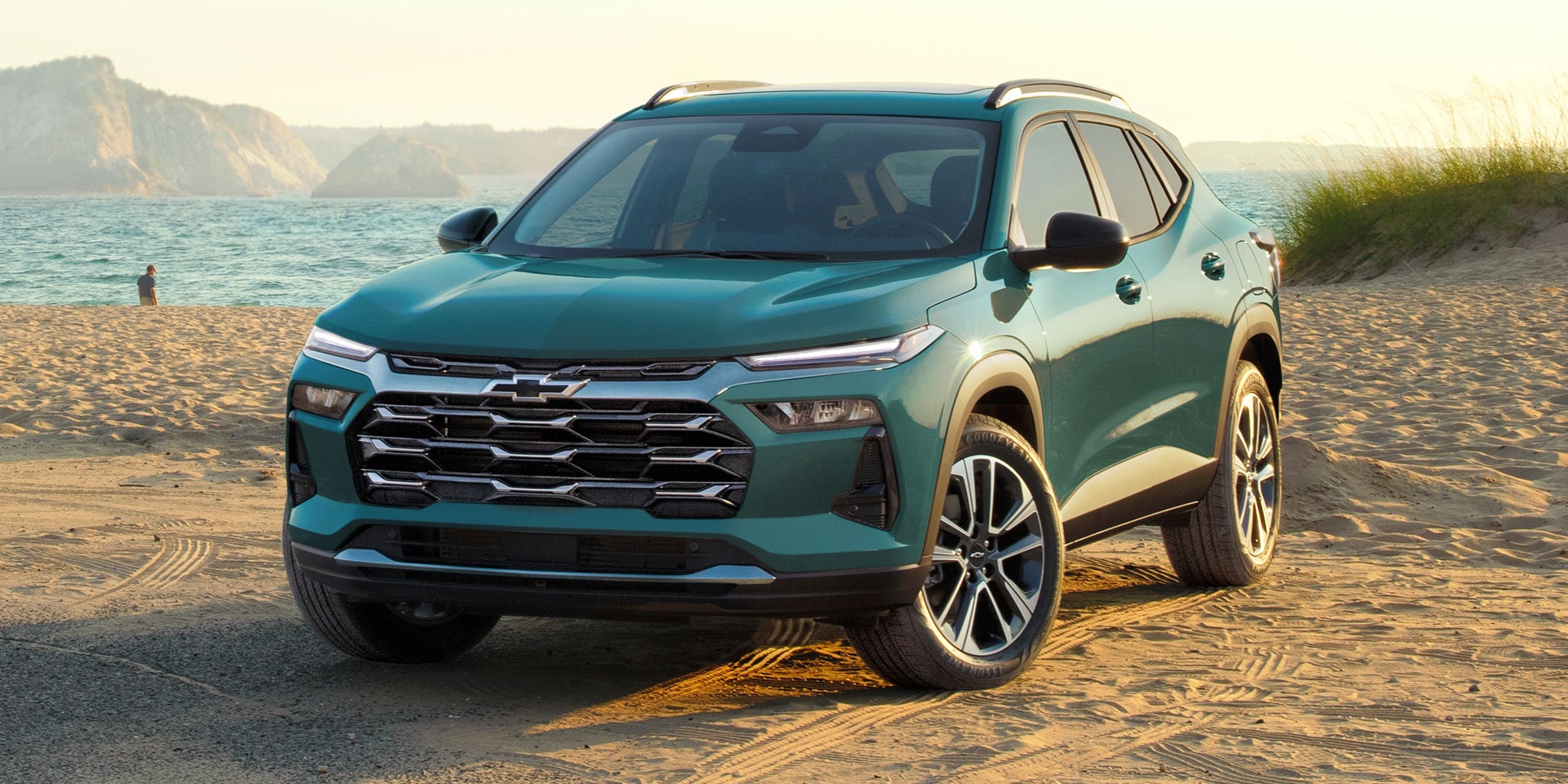 Green 2027 Chevrolet Trax small SUV parked on a sandy beach, shown from a front three-quarter angle with ocean water, coastal dunes, and distant cliffs in the background.