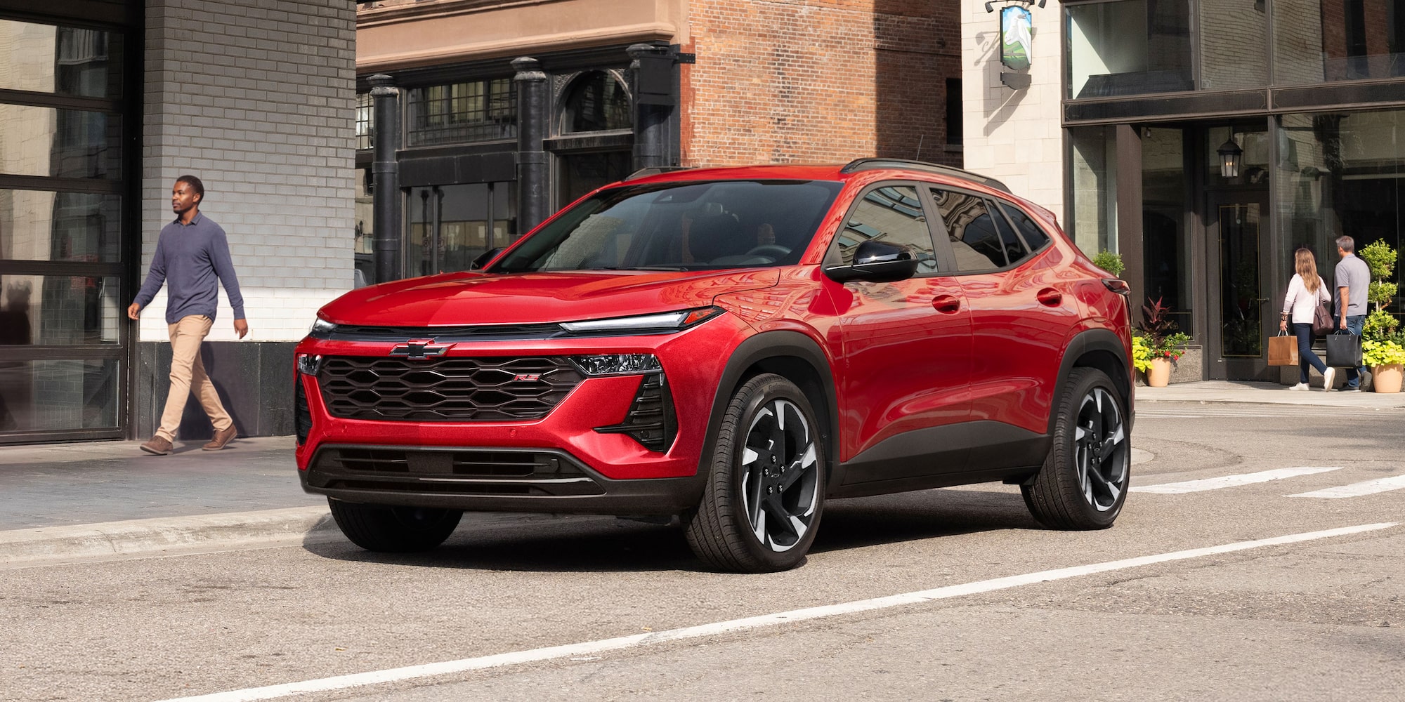 Red 2027 Chevrolet Trax small SUV parked on a city street, shown from a front three-quarter angle with modern urban buildings and storefronts in the background.