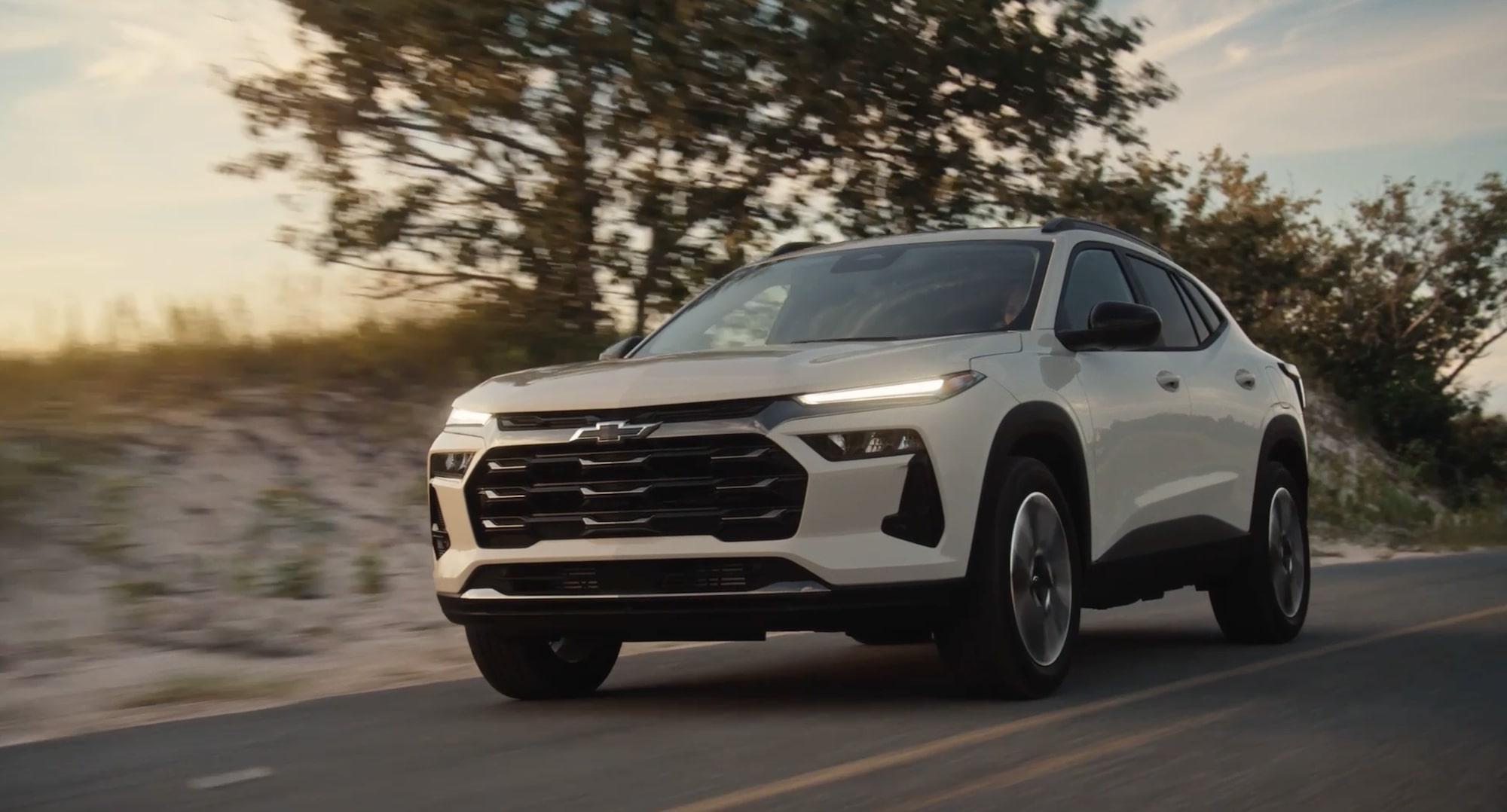 White 2027 Chevrolet Trax small SUV driving on a paved road, shown from a front three-quarter angle with trees and sandy dunes in the background.