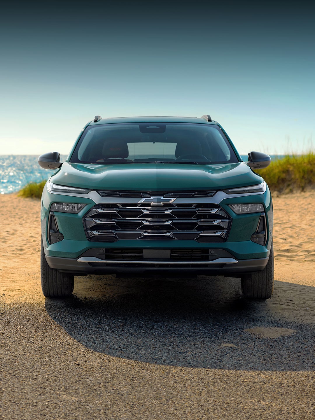 Front three-quarter view of a green 2027 Chevrolet Trax small SUV parked on a sandy beach with ocean water and coastal rock formations in the background.